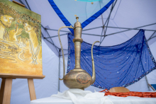 A metal teapot with a long spout and decorative details stands on a table. Next to it, there is a painting on an easel as well as fabrics and decorations. Everything is under a tent.
