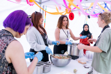 Five people stand at a table preparing food. One person serves a portion from a pot. A bowl of salad and food containers are on the table.