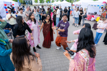 A group of about 10 people stands in a circle and dances. They raise their hands and move to the rhythm of the music. Stalls and event participants are visible in the background.