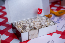 An open cardboard box stands on a table and contains several dozen pieces of a sweet snack dusted with powdered sugar. Inside, there is a small Turkish flag on a stick. The table is covered with a red-and-white checkered cloth.