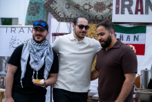 Three people stand side by side at a stall and pose for a photo. One person is holding a plate of food. Behind them hang decorative fabrics and the Iranian flag. Elements of the stall and tableware are visible in the background.