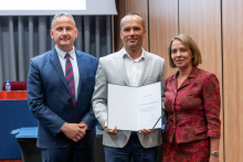 Two men and a woman are posing together for a photo. The man in the middle is holding an open diploma in his hands.