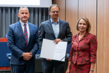 Two men and a woman are posing together for a photo. The man in the middle is holding an open diploma in his hands.