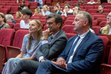 Three audience members sit in the front row of a lecture hall with red seats, facing the stage. One person holds a folder on their lap, and all are looking forward.