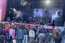 A wide view of a live music concert with a band performing on a large outdoor stage. A crowd of people is gathered in front of the stage, watching the performance, with screens and stage lighting visible above the musicians.