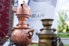 Two metal teapots stand in the foreground. Behind them, there is a poster with the words “Welcome to IRAN.” Patterned fabrics and decorative elements are visible nearby.