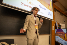Person standing in front of a projection screen and holding a handheld microphone while speaking. Logos and event information are visible on the screen behind.