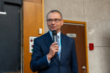 One person in a suit stands by a wall and holds a microphone while speaking; doors and room furnishings are visible in the background.