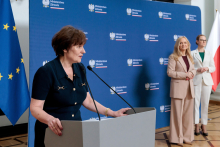 One person speaks at a lectern with microphones, holding the edge of the podium with both hands. Behind this person, two individuals stand side by side, one holding a document. The background features a blue wall with “Ministerstwo Zdrowia” text and a European Union flag.