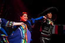 Three performers dancing on stage, wearing traditional costumes, with one person holding a wide-brimmed hat during a choreographed movement.