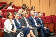 An audience seated in a lecture hall with red chairs, where several attendees in formal attire sit in the front row, some holding eyeglasses or resting hands on their laps, while additional participants are seated in the rows behind.