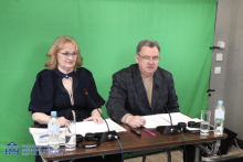 Two people seated behind a desk with microphones, documents, and bottles of water placed in front of them. Headphones lie on the table. A green screen covers the entire background, indicating a recording or broadcasting setup.