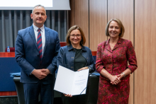 A man and two women are posing together for a photo. The woman in the middle is holding an open diploma in her hands.
