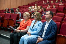 Front row of audience members seated in red chairs. One person is holding a printed program, while others are sitting with hands folded, facing the stage