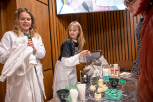A workshop station with educational materials. People in protective lab coats stand by a table holding tooth models, silicone molds, bowls, tools, and substances for practical activities.