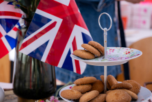 A two-tiered stand sits on a table and holds several cookies. In the background, the flag of the United Kingdom and a glass pitcher can be seen.