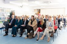 Participants are seated in rows of chairs in a bright conference room. People in the front row hold notebooks and documents in their hands.
