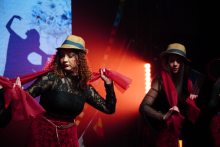 Two performers on stage wearing hats and red scarves, moving in unison under warm stage lights.