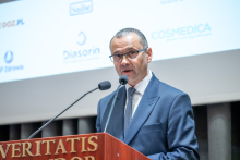 A person in a dark blue suit and glasses stands at a lectern with two microphones, delivering a speech. The lectern displays the inscription “VERITATIS SPLENDOR.” In the background, a projection screen shows logos including “