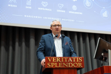 A person standing at a lectern with the inscription ‘Veritatis Splendor’, speaking into microphones. In the background, a projection screen displays conference graphics and the name of the Medical University of Warsaw