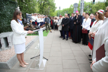 Osoba przy mównicy czyta z przygotowanego tekstu, trzymając kartki w dłoniach. Przed nią stoją uczestnicy uroczystości, część trzyma teczki lub dokumenty. Obok znajduje się pomnik oraz kamienny element memorialny.