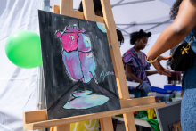 An easel displays a painting of a simplified heart silhouette in shades of pink and blue. Two people stand next to it at a table preparing items.