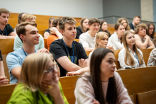 Audience sitting on wooden benches in a lecture hall and looking toward the front of the room. Some people are wearing lanyards.