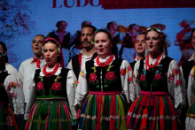 A group of performers standing in a row on stage, wearing matching traditional embroidered costumes with colorful skirts.