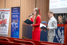 Two people stand at the front of a conference room holding microphones and folders. One person in a red outfit reads from an open folder while the other in a light grey suit holds a closed folder and a microphone. Behind them are banners and a screen with logos, including “Pharmaceris,” and a lectern with an emblem.