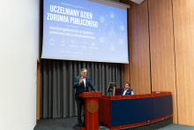 A wide view of a lecture hall stage where a person speaks at a lectern beneath a screen reading ‘University Public Health Day’. Another seated person is visible behind a desk on the stage.
