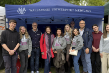 A group of people standing together in front of a blue canopy tent labeled “Medical University of Warsaw”. The group is posing outdoors, with trees and event tents visible in the background.