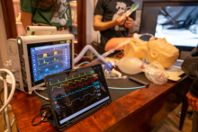 A demonstration table with medical equipment. In the foreground, monitor screens displaying vital parameter charts are visible, alongside a medical mannequin with a ventilation mask and training equipment elements.