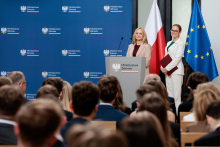 Dwie osoby stoją przy mównicy z godłem Rzeczypospolitej Polskiej. Jedna osoba przemawia, druga stoi obok i trzyma dokumenty. Przed mównicą siedzi publiczność. W tle znajdują się flagi Unii Europejskiej i Polski oraz ścianka z logotypami Ministerstwa Zdrowia.