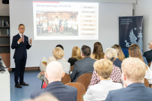 A speaker stands by a screen and delivers a presentation, gesturing with their hands. The screen displays the text “Department of Cardiology, UCK WUM,” and the audience is seated, with some people holding materials and a bouquet of flowers.