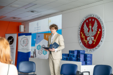 One person stands in a conference room holding a folder; boxes with the ALAB logo are placed nearby, with the university emblem and standing banners in the background.