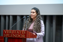 A person with long hair stands behind a lectern with two microphones, speaking during a presentation. The lectern bears the inscription “VERITATIS SPLENDOR.” A projection screen is visible in the background.