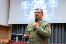 A person in a formal uniform giving a presentation while holding a microphone. Behind the speaker, a presentation slide with icons and diagrams related to health and safety is displayed