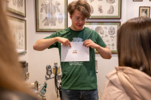 An educational presentation in a museum room. A person standing in the center shows a sheet with an illustration, surrounded by participants; anatomical charts and medical devices are visible in the background.
