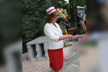 Osoba w jasnym stroju i czepku pielęgniarskim stoi przy mównicy, trzymając dokument i przemawiając do zgromadzonych. Za mównicą widoczny jest pomnik z biało‑czerwoną wstęgą oraz osoba z aparatem fotograficznym trzymanym przy twarzy