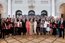 Grupowe ujęcie kilkudziesięciu osób stojących w kilku rzędach w reprezentacyjnej sali. Uczestnicy trzymają bordowe teczki z godłem. W centrum stoją osoby uczestniczące w części oficjalnej wydarzenia. W tle widoczne są arkady, kolumny oraz elementy wystroju sali.