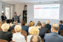 A presenter stands by the screen and points to a slide. The audience sits in rows and looks at the screen.