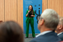 A person standing on a stage and speaking into a handheld microphone. Behind the speaker, a blue banner displays the Medical University of Warsaw Faculty of Health Sciences logo. Audience members are visible in the foreground.