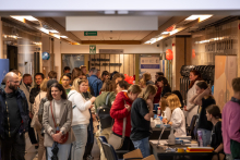 A crowded museum corridor with numerous educational stands. Visitors stand at workshop tables, talk to instructors, and examine educational materials; balloons and elements of the permanent exhibition are visible in the background.