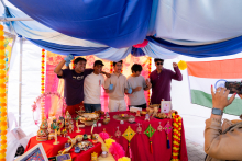 Five people stand behind a table with decorations and food. One person is taking a photo with a phone. Decorative items and snacks are placed on the table.