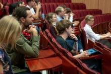 Audience members sit in tiered red seats in a lecture hall, facing the front. One person uses a tablet placed on the fold-out writing surface, while others sit with hands clasped.