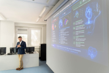 A presenter stands next to a large screen with a medical presentation and holds a slide remote in their hand.