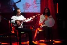 Two seated musicians on stage, one playing a string instrument and the other a frame drum, under red stage lighting.