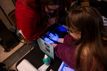 An educational station with a UV lamp. A child puts their hands into a device emitting blue light, and hand hygiene disinfectants and informational materials are placed on the table nearby.