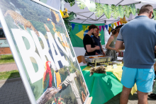 A large poster with the word “BRAZIL” stands next to a table. Three people stand nearby, running a food stall. The table holds dishes and tableware.