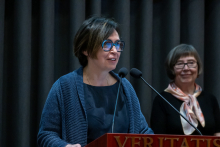 Person speaking at a podium with microphones. Another person stands in the background near the same podium.
